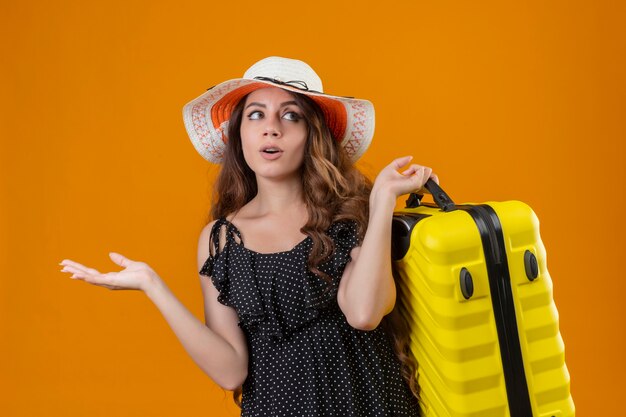 Junges schönes reisendes Mädchen im Kleid im Tupfen im Sommerhut, der den ahnungslosen und verwirrten Koffer hält, der Zweifel hat, die über gelbem Hintergrund stehen