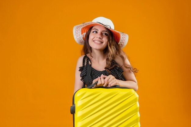 Junges schönes reisendes Mädchen im Kleid im Tupfen im Sommerhut, der Koffer hält, der beiseite schaut, fröhlich glücklich und positiv stehend über orange Hintergrund