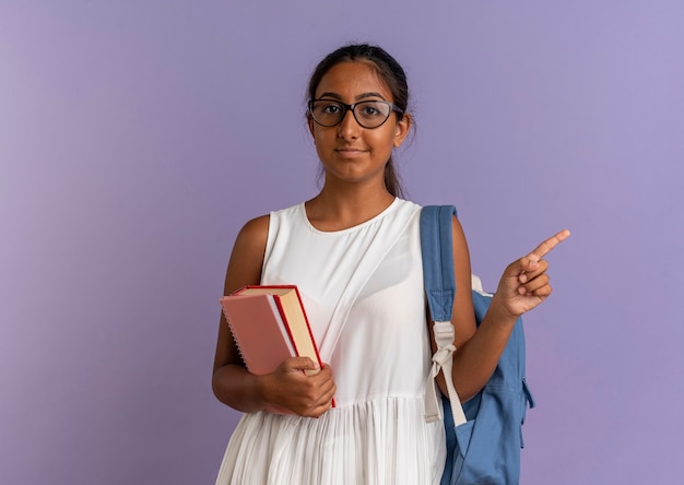 junges Schulmädchen, das Rucksack und Brille hält, Bücher hält und zur Seite zeigt
