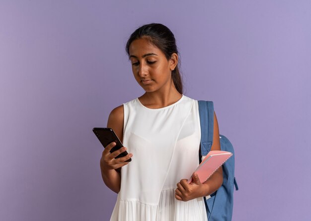 Junges Schulmädchen, das Rückentasche hält, das Notizbuch hält und Telefon in ihrer Hand auf lila betrachtet