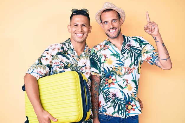Junges schwules Paar von zwei Männern, die einen Koffer in den Sommerferien halten, überrascht mit einer Idee oder Frage, die mit dem Finger mit glücklichem Gesicht, Nummer eins, zeigt