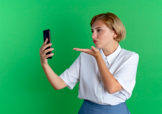 junges selbstbewusstes blondes russisches Mädchen hält und schickt Kuss mit der Hand zum Telefon