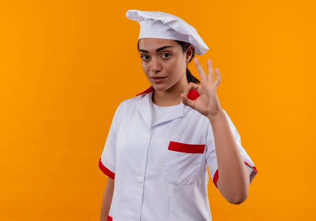 Junges selbstbewusstes kaukasisches Kochmädchen in der Kochuniform gestikuliert ok Handzeichen lokalisiert auf orange Wand mit Kopienraum