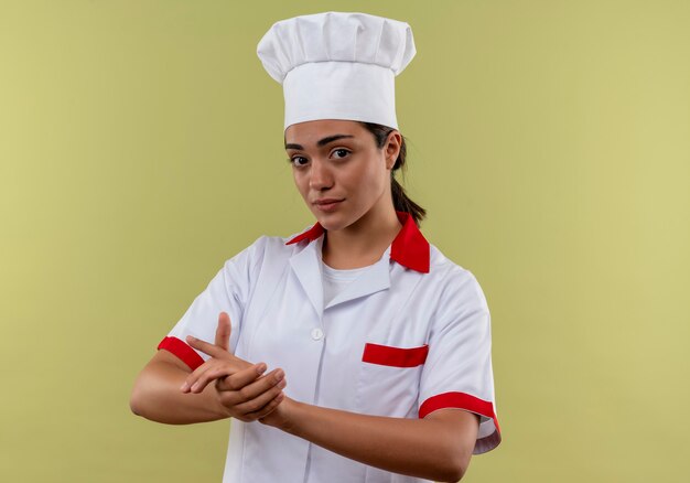 Junges selbstbewusstes kaukasisches Kochmädchen in der Kochuniform hält Hände zusammen lokalisiert auf grüner Wand mit Kopienraum