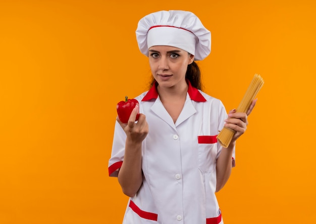 Junges selbstbewusstes kaukasisches Kochmädchen in der Kochuniform hält roten Pfeffer und Bündel Spaghetti lokalisiert auf orange Wand mit Kopienraum