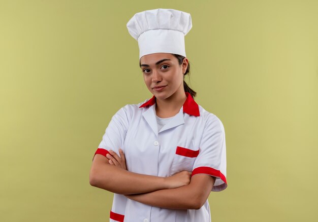 Junges selbstbewusstes kaukasisches Kochmädchen in der Kochuniform kreuzt Arme und betrachtet Kamera lokalisiert auf grünem Hintergrund mit Kopienraum