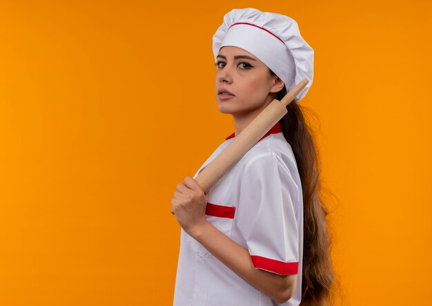 Junges selbstbewusstes kaukasisches Kochmädchen in der Kochuniform steht seitlich und hält Nudelholz isoliert auf orange Wand mit Kopienraum