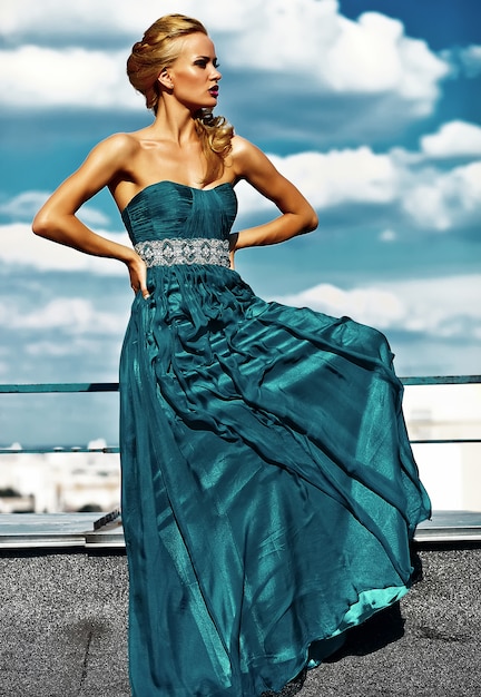 Junges sexy blondes Frauenmodell im Abendkleid, das auf Hintergrund des blauen Himmels aufwirft