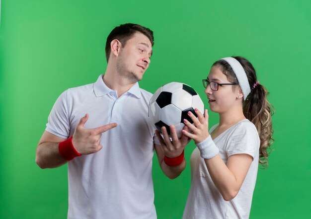Junges sportliches Paar Mann und Frau, die nebeneinander stehen und Fußball über grüner Wand halten