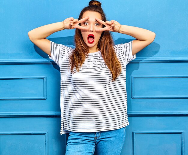 Junges stilvolles Frauenmodell im zufälligen Sommer kleidet mit den roten Lippen und wirft nahe blauer Wand auf. Überrascht und zeigt friedenszeichen