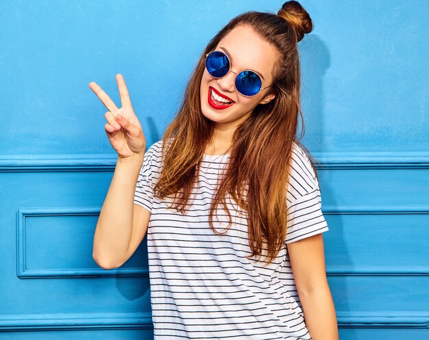 Junges stilvolles Frauenmodell im zufälligen Sommer kleidet mit den roten Lippen und wirft nahe blauer Wand auf. Zeige Friedenszeichen