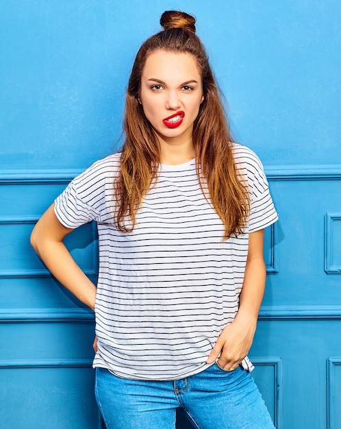 Junges stilvolles Frauenmodell im zufälligen Sommer kleidet mit den roten Lippen und wirft nahe blauer Wand auf