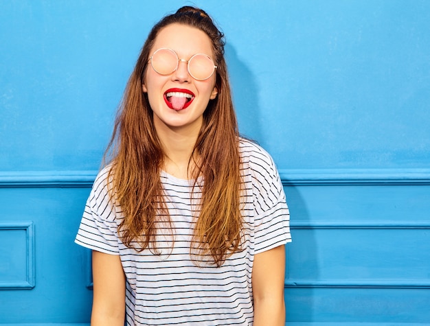 Kostenloses Foto junges stilvolles frauenmodell im zufälligen sommer kleidet mit den roten lippen und wirft nahe blauer wand, in der sonnenbrille auf. zeigt ihre zunge