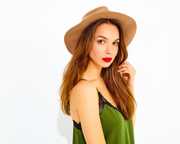Junges stilvolles Frauenmodell in der zufälligen Sommergrünkleidung und im braunen Hut mit den roten Lippen, werfend nahe weißer Wand auf
