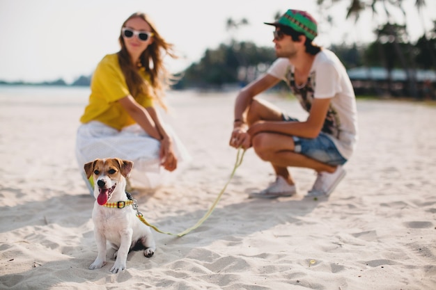 Kostenloses Foto junges stilvolles hipster-paar in der liebe, das das spielen des hundewelpen jack russell im tropischen strand, im weißen sand, im kühlen outfit, in der romantischen stimmung spielt, spaß, sonnig, mann frau zusammen, urlaub