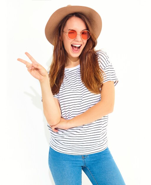Junges stilvolles lachendes Frauenmodell im zufälligen Sommer kleidet im braunen Hut mit dem natürlichen Make-up, das auf weißer Wand lokalisiert wird. Friedenszeichen blinzeln und zeigend