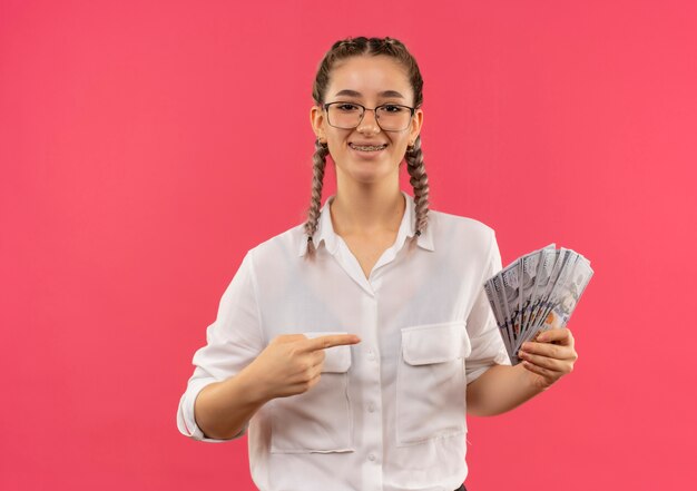 Junges Studentenmädchen in den Gläsern mit Zöpfen im weißen Hemd, das Bargeld hält, das mit dem Finger auf es zeigt, das zuversichtlich steht, über rosa Wand zu stehen