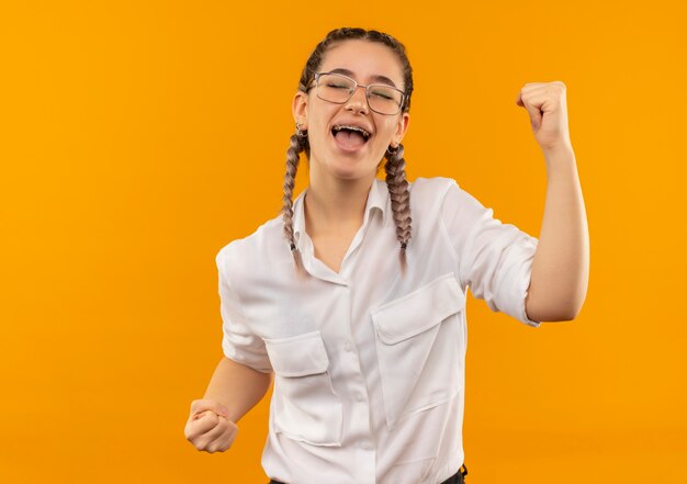 Junges Studentenmädchen in den Gläsern mit Zöpfen im weißen Hemd, das die Fäuste glücklich und aufgeregt ballt, die ihren Erfolg freuen, der über orange Hintergrund steht