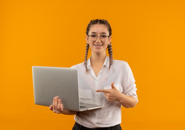 Junges Studentenmädchen in den Gläsern mit Zöpfen im weißen Hemd, das Laptop hält, der mit dem Finger auf ihn lächelt, der zuversichtlich über orange Wand steht