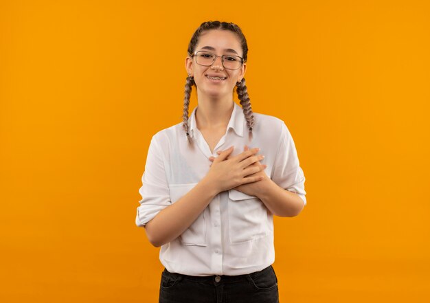Junges Studentenmädchen in den Gläsern mit Zöpfen im weißen Hemd, das nach vorne mit Händen schaut, die auf der Brust lächelnd fühlen und dankbar sind, über orange Wand stehend