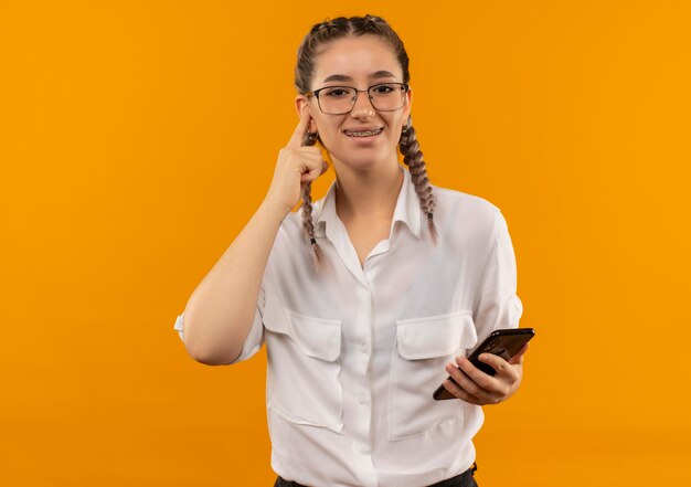 Junges Studentenmädchen in den Gläsern mit Zöpfen im weißen Hemd, das Smartphone hält, das mit Finger zum Tempel zeigt, der über orange Wand lächelt