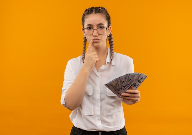 Junges Studentenmädchen in Gläsern mit Zöpfen im weißen Hemd, das Bargeld hält, das nach vorne mit nachdenklichem Ausdruck denkt, der Zweifel hat, die über orange Wand stehen