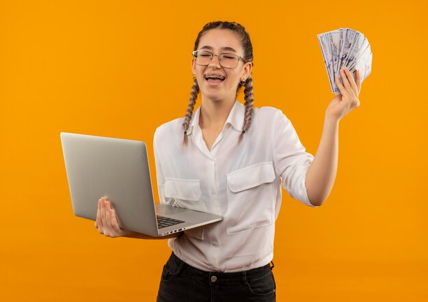 Junges Studentenmädchen in Gläsern mit Zöpfen im weißen Hemd, das Laptop und Bargeld glücklich und aufgeregt lächelt, das fröhlich über orange Wand steht