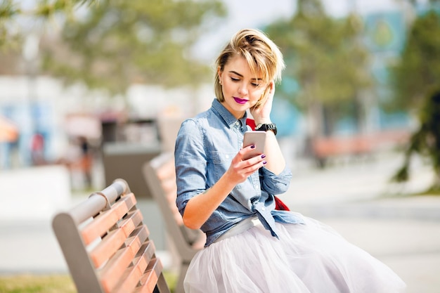 Junges süßes blondes Mädchen mit kurzen Haaren, das auf einer Holzbank sitzt und das Smartphone mit blauem Jeanshemd und grauem Tüllrock betrachtet, das ihr Haar hinter dem Ohr kämmt.