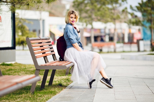 Junges süßes blondes Mädchen mit kurzen Haaren und leuchtend rosa Lippen, das auf einer Holzbank sitzt und lächelt, trägt blaues Jeanshemd, grauen Tüllrock, schwarze Turnschuhe und Marsala-Rucksack.