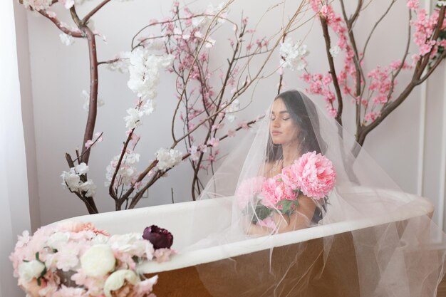 Junges süßes Model sitzt in einem Bad unter dem weißen Tüll mit Blumen Hochwertiges Foto