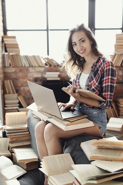Junges Teenager-Mädchen, das den Laptop-Computer verwendet, der durch viele Bücher umgeben wird.