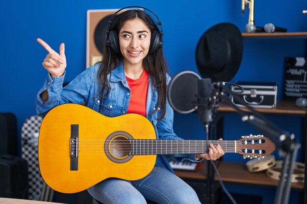Junges Teenager-Mädchen, das im Musikstudio klassische Gitarre spielt und mit dem Daumen zur Seite zeigt, glücklich lächelnd und mit offenem Mund