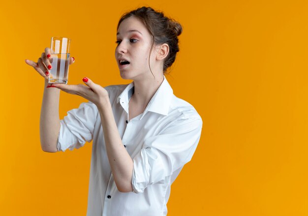 Junges überraschtes blondes russisches Mädchen hält und zeigt auf Glas Wasser auf Orange mit Kopienraum