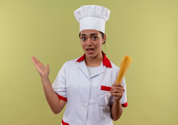 Junges überraschtes kaukasisches Kochmädchen in der Kochuniform hält Bündel Spaghetti und hebt Hand lokalisiert auf grüner Wand mit Kopienraum auf