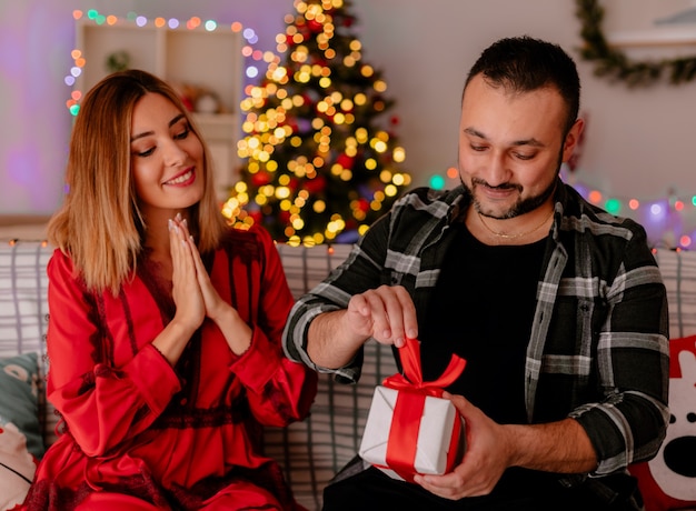 junges und schönes Paar, das auf einem Couch-Mann sitzt, der ein Geschenk öffnet, während seine glückliche Freundin ihn betrachtet, die Weihnachten zusammen im geschmückten Raum mit Weihnachtsbaum im Hintergrund feiert