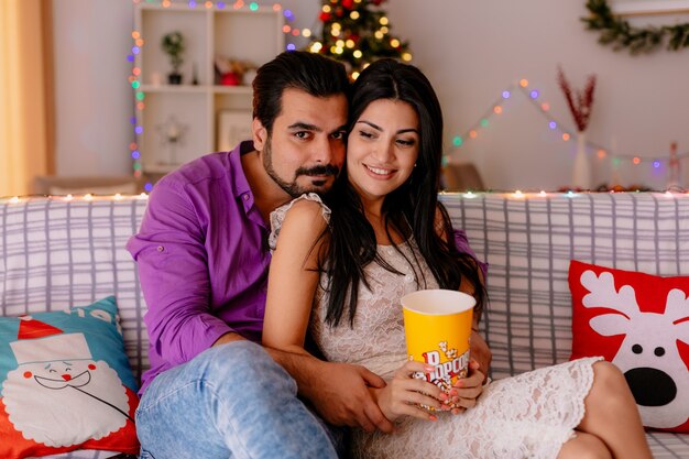 junges und schönes Paar, das auf einer Couch Mann und Frau mit Eimer Popcorn sitzt zusammen fernsehen glücklich in der Liebe in dekoriertem Raum mit Weihnachtsbaum im Hintergrund