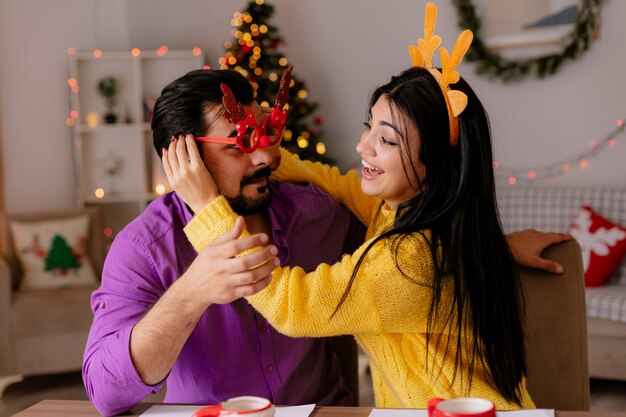 Junges und schönes Paar Mann und Frau, die am Tisch mit Keksen sitzen, die Spaß zusammen glücklich in der Liebe im weihnachtlich geschmückten Raum mit Weihnachtsbaum im Hintergrund haben