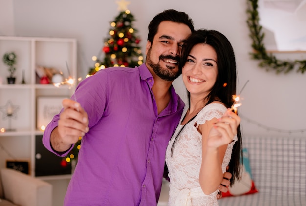 junges und schönes Paar Mann und Frau mit Wunderkerzen umarmt glücklich in der Liebe, die Weihnachten zusammen im geschmückten Raum mit Weihnachtsbaum im Hintergrund feiert