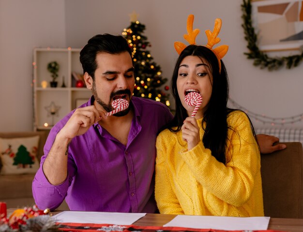 junges und schönes Paar Mann und Frau mit Zuckerstangen, die Spaß zusammen glücklich in der Liebe in weihnachtlich geschmücktem Raum mit Weihnachtsbaum im Hintergrund haben