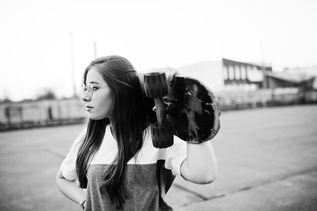 Junges urbanes Mädchen im Teenageralter mit Skateboardabnutzung auf Brillenkappe und zerrissenen Jeans auf dem Hofsportplatz bei Sonnenuntergang