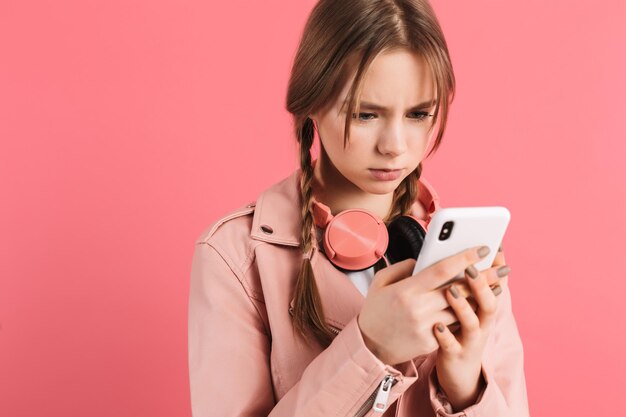 Junges verärgertes Mädchen mit zwei Zöpfen in Lederjacke mit Kopfhörern, das traurig Handy benutzt, während es Zeit auf rosafarbenem Hintergrund verbringt