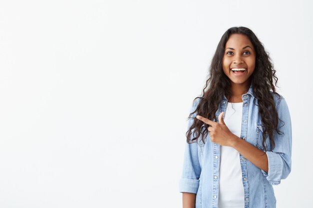 Junges verblüfftes afroamerikanisches Mädchen mit langem gewelltem Haar, das mit geöffnetem Mund Zähne zeigt und ihren Finger auf weiße Wand mit Kopienraum für Ihre Werbung oder Verkaufsförderung zeigt