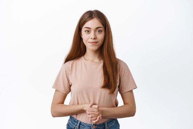Junges weibliches Modell, das bereit zu Ihren Diensten steht und Händchen hält, das sich in der Nähe der Brust zusammenhält, um zu helfen, lächelnd und entschlossen auf weißem Hintergrund zu stehen