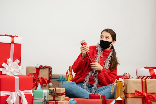 junges Weihnachtsmädchen mit schwarzer Maske, die Party-Popper hält, der herum Geschenke auf Weiß sitzt