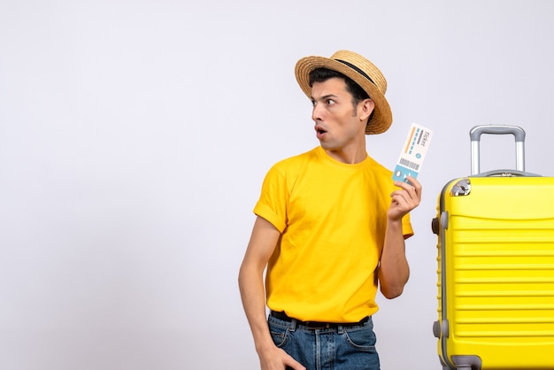 Jungtourist der Vorderansicht im gelben T-Shirt, das nahe gelbem Koffer steht, der links hält Ticket betrachtet