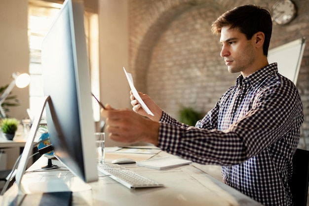 Jungunternehmer, der Computer verwendet, während er Daten mit Geschäftsberichten im Büro vergleicht