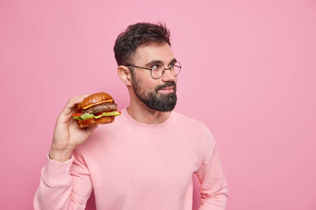 Junk-Food und ungesundes Ernährungskonzept. Bärtiger erwachsener Mann hält leckeren Hamburger hat schnellen Snack schaut nachdenklich weg away