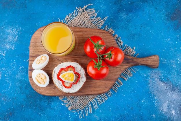 Käsebrot, ein Glas Saft, geschnittenes Ei und ganze Tomaten auf Schneidebrett auf Stoffstücken auf Blau.