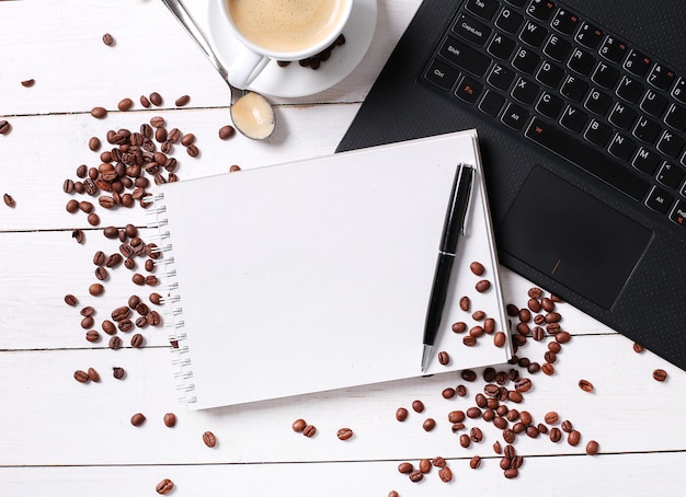 Kaffeebohnen auf leerem Notizbuch, Kaffeetasse und Laptop