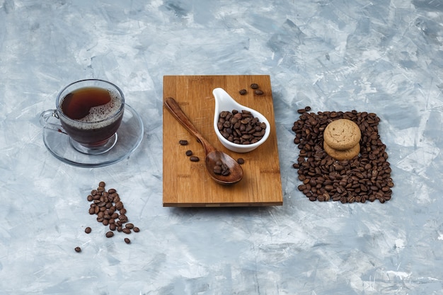 Kaffeebohnen mit hohem Blickwinkel, Holzlöffel auf Schneidebrett mit Keksen, Tasse Kaffee auf hellblauem Marmorhintergrund. horizontal